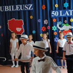 Scenka z przedstawienia - w tle śpiewające dziewczynki, na pierwszym planie chłopcy w kapeluszach, z parasolami.