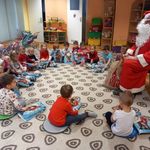 Mikołaj siedzi na krześle,a wokół niego zgromadziły się dzieci, które śpiewają jemu piosenki.